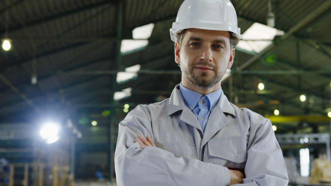 Caucasian engineer wearing a helmet turning his head to the camera, smiling and crossing arms in a factory