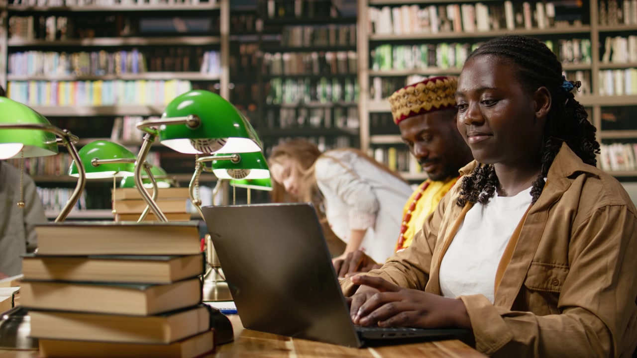 Black woman student uses laptop at shared library table with stacked books