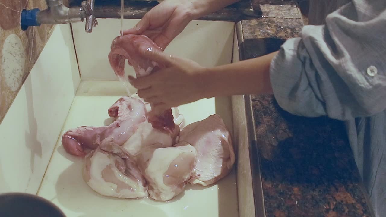 Person washing raw pork tenderloin and poultry under a faucet in a kitchen sink that shows daily home chores and candid domestic life