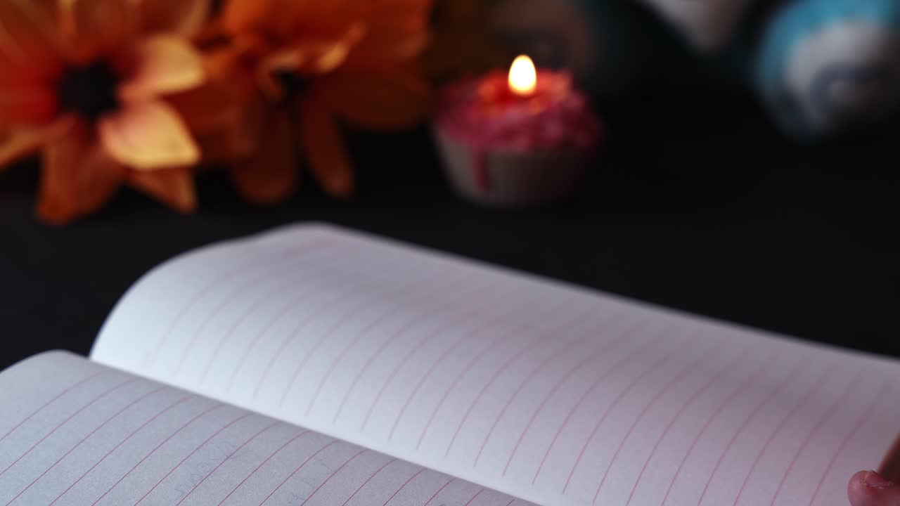 Close-up of open lined notebook with handwritten romantic message “I love you” and heart doodle, hand holding page, warm candlelight and flower in soft indoor background
