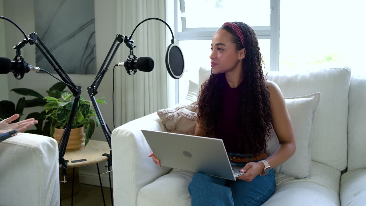 Woman in burgundy touching hair using laptop gesturing to cohost on couch with mic-booms recording