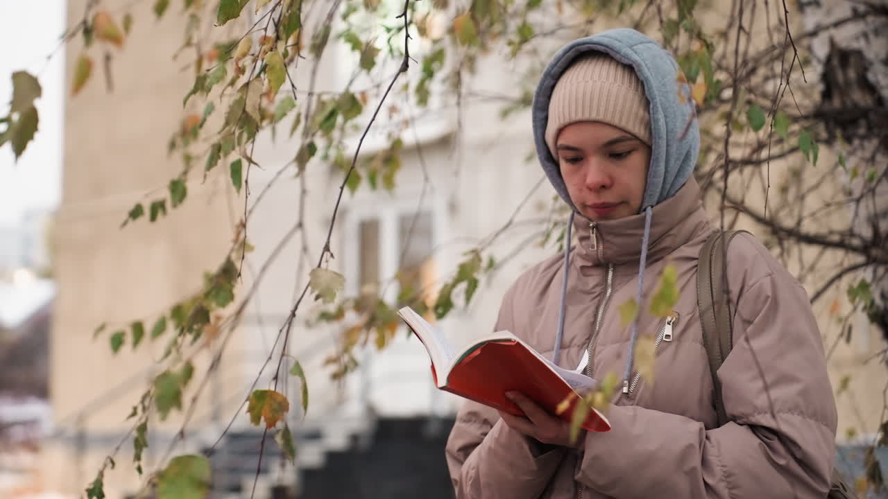 Light skin lady dressed in beige winter coat and beanie stands beneath tree with yellowing leaves in autumn, flipping through red book pages with concentration, blurred residential building in background