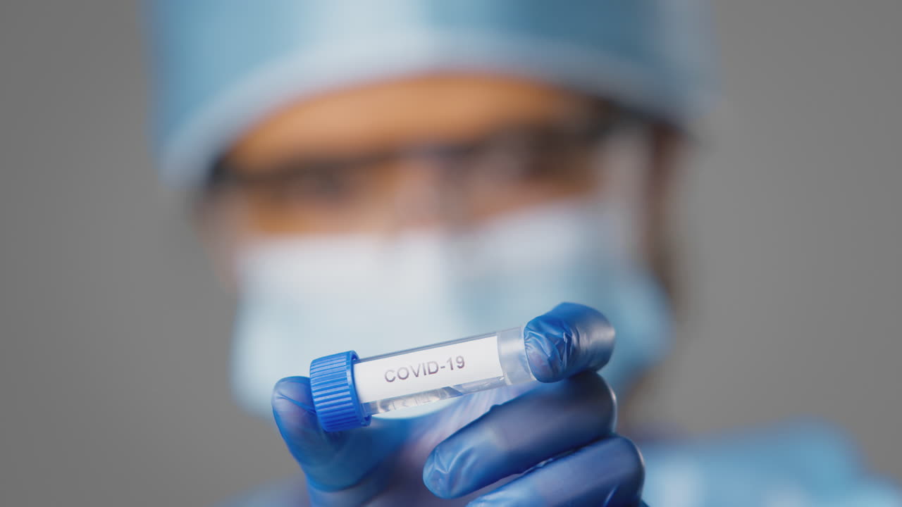Primo piano di una ricercatrice di laboratorio che indossa DPI e tiene una provetta etichettata Covid 19