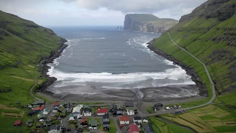 aerial view of tjørnuvík village, nestled between lush green cliffs and the ocean in the faroe islands. features black sand beach, vibrant houses, and striking coastal scenery with cliffs and waves