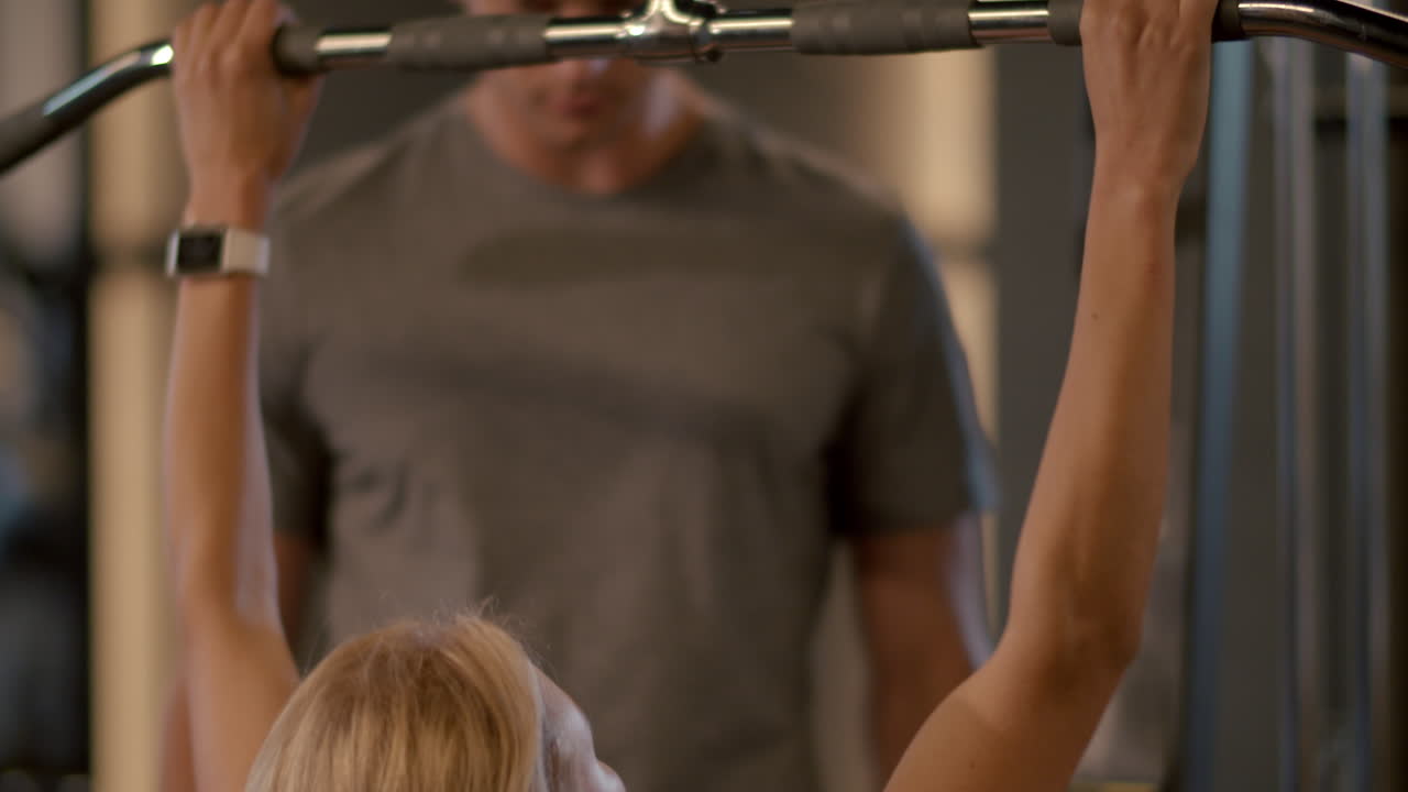 Serious fit girl making pulldown in sport club. Trainer controlling client