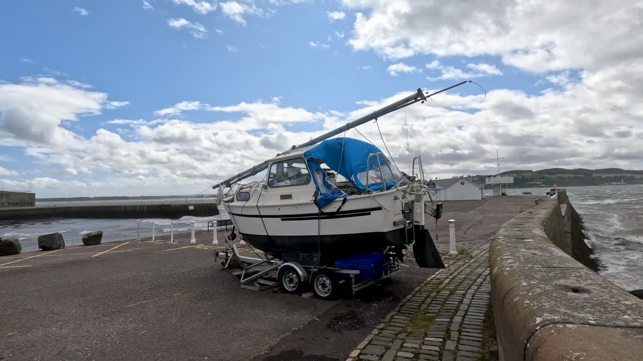 A Hardy motor sailer boat on a trailer is moved along a coastal promenade at Broughty Ferry, Dundee, under partly cloudy daylight with steady camera panning