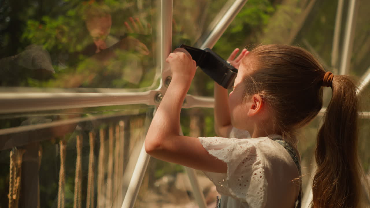 niño con cámara digital en tienda de glamping. niña pequeña dispara belleza del paisaje a través de la pared de vidrio en un día soleado. niño toma fotos del campamento