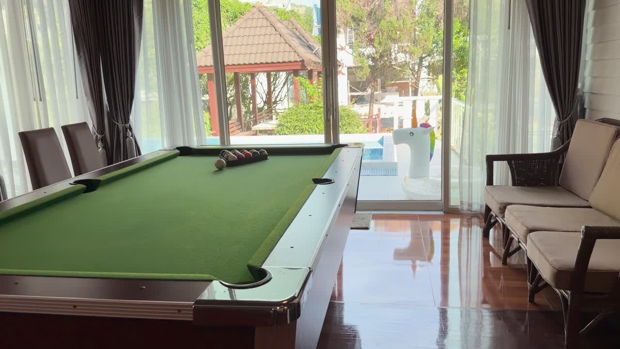 Camera pans from an indoor pool table toward large windows overlooking a sunny swimming pool