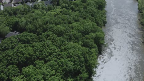 Drohne-Fliegt-über-Einen-Fließenden-Fluss-Im-Mittleren-Westen-Der-USA,-Schwenkt-Nach-Oben-Und-Zeigt-Eine-Vorstädtische-Wohnsiedlung-Am-Flussufer