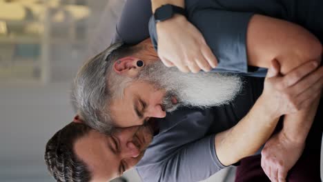 Vertical-video-of-a-sad-man-looking-for-support-and-leaning-on-his-brunette-boyfriend-with-stubble-in-a-gray-T-shirt-while-sitting-on-the-sofa-in-a-modern-apartment.-Elderly-man-leans-on-his-boyfriend-during-sad-mood-LGBT-couple