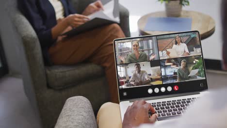 hombre afroamericano usando una computadora portátil para una llamada de video, con diversos colegas de negocios en la pantalla