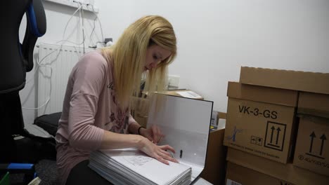 una chica de pelo rubio sentada en el suelo clasificando a través de un gran archivo de documentos al lado de una pila de cajas de cartón, preparándose para mudarse de un apartamento