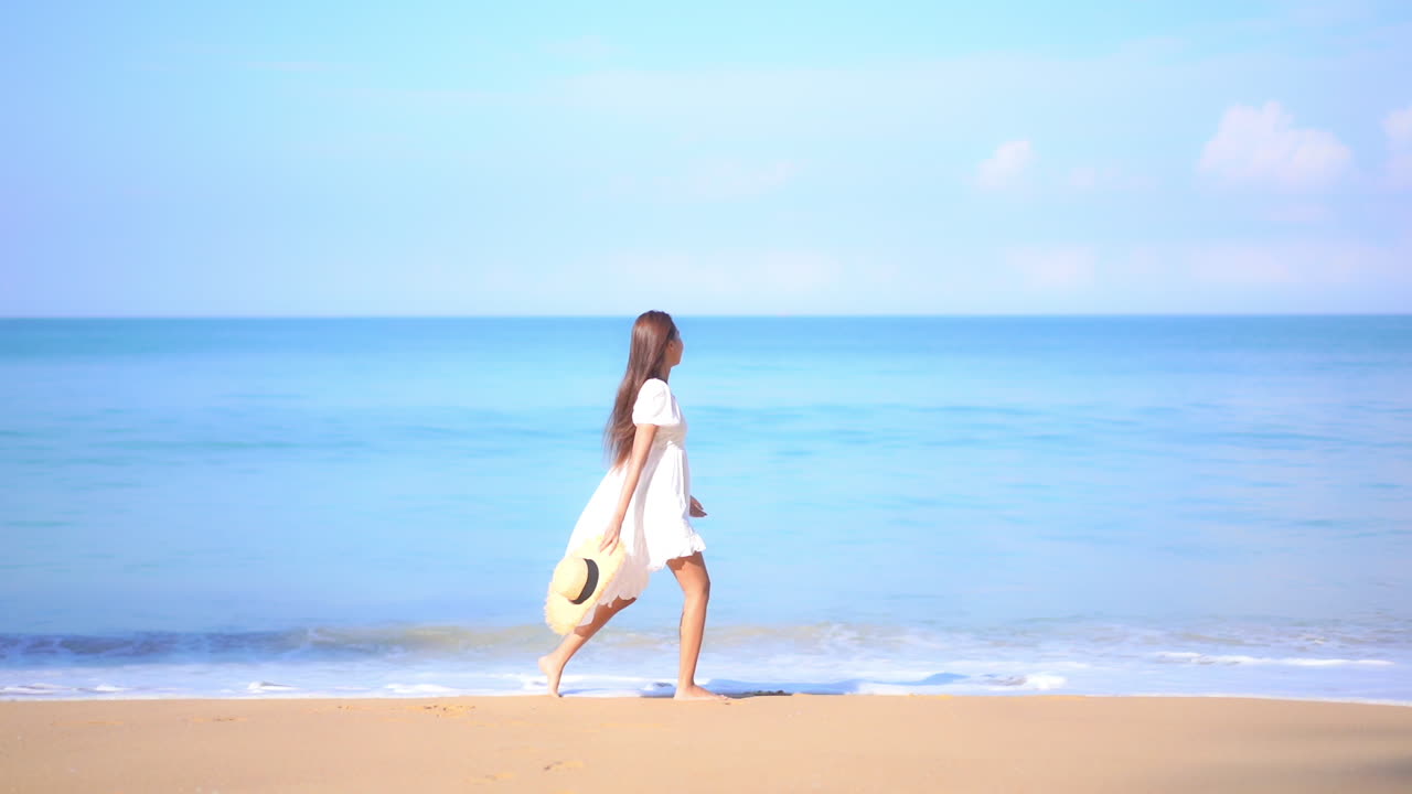 열대 바다 앞의 모래사장을 손에 들고 걷고 있는 하얀 드레스를 입은 매력적인 여성, 슬로우 모션