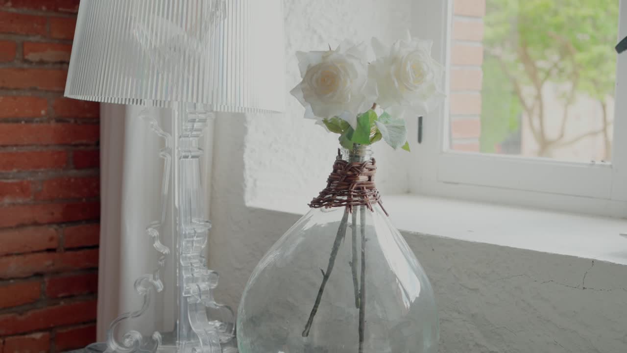 elegantes rosas blancas en un jarrón de vidrio en un alféizar de la ventana, junto a una lámpara transparente, creando una decoración serena y elegante