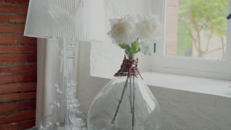 elegant white roses in a glass vase on a windowsill, next to a transparent lamp, creating a serene and stylish decor