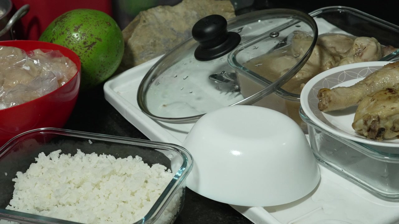Prepared white rice, cooked chicken drumsticks, and other leftovers rest in glass containers on a busy countertop tray, suggesting home meal storage after cooking.