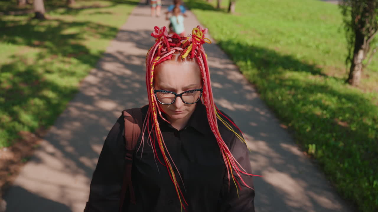 artista solitario de piel blanca paseando por un parque, rastas de color rojo brillante y amarillo recogidas, gafas de gran tamaño, paso lento y deliberado por una avenida frondosa, luz moteada suave, reflexión interior y pausa emocional