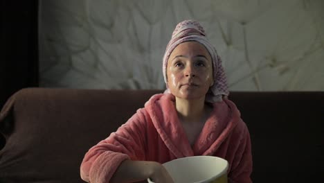 Mujer-Viendo-Una-Película-Nocturna-En-La-Televisión,-Comiendo-Palomitas-De-Maíz.-Bata-De-Baño,-Mascarilla-Facial