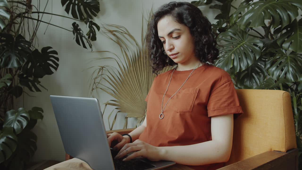 chica biracial en un sillón y usando una computadora portátil en una habitación con plantas