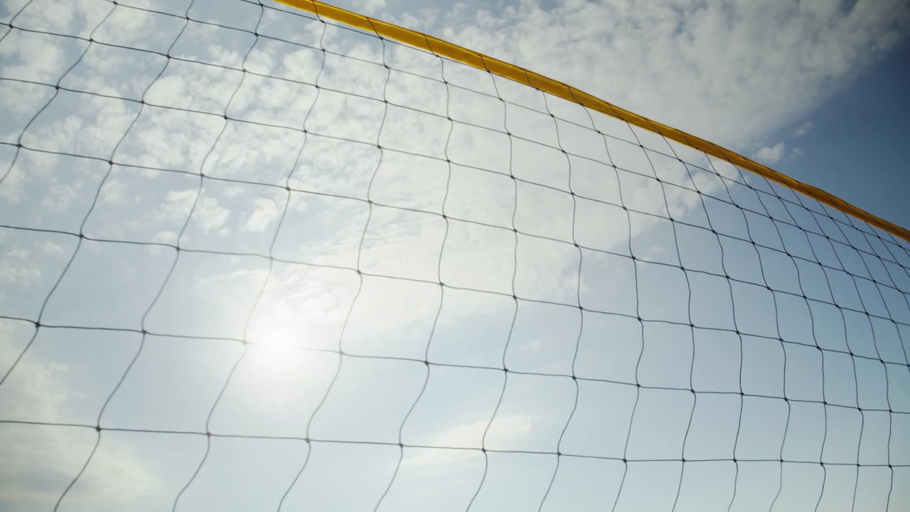 Pemandangan dekat jaring bola voli pantai dengan latar belakang langit biru cerah, awan putih, dan matahari bersinar di hari musim panas