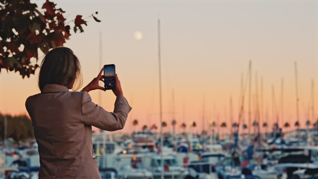Rear view of a woman taking a smartphone photo of a marina full of boats at sunset, with the moon and palm trees in the soft pastel sky