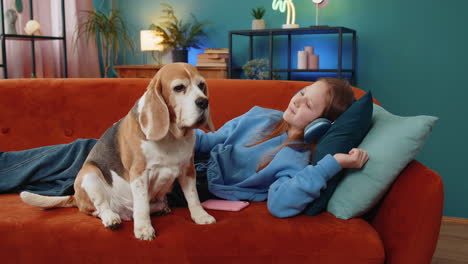 child girl kid in wireless headphones relaxing listening music lying on sofa at home near beagle dog