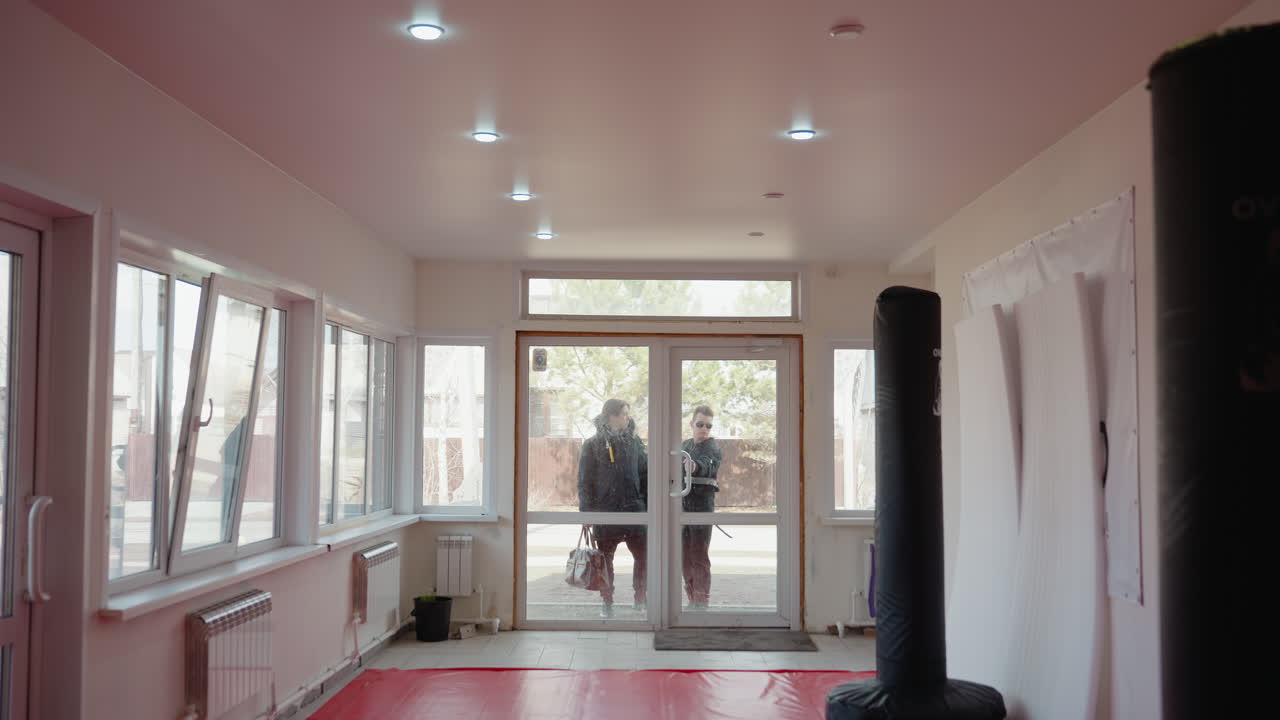 Two guys in jackets approach glass entrance of training hall, one carrying leather bag, viewed from inside room with punching bags, mats, radiators and windows, urban lifestyle moment in daylight atmosphere