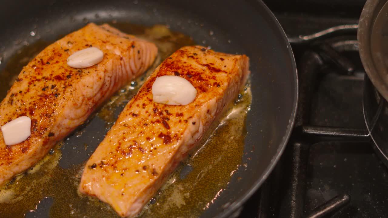 Overhead View of Two Salmon Fillets Cooking in Frying Pan with Butter Gently Bubbling and Garlic Cloves on Top for Additional Flavour. Slow Motion Footage 4K