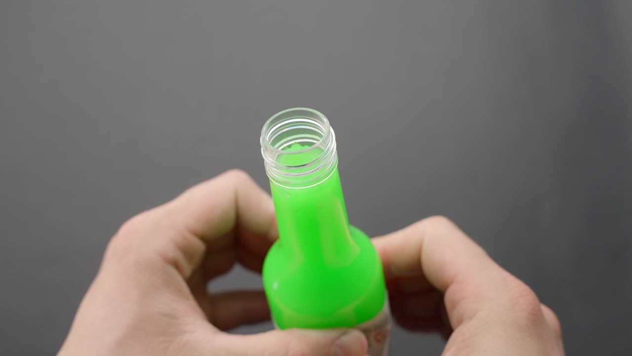 Close-up of hands pressing vibrant green floam slime through the neck of a clear bottle, creating playful texture and squishy effect, set against plain grey background, focus on tactile sensation ASMR