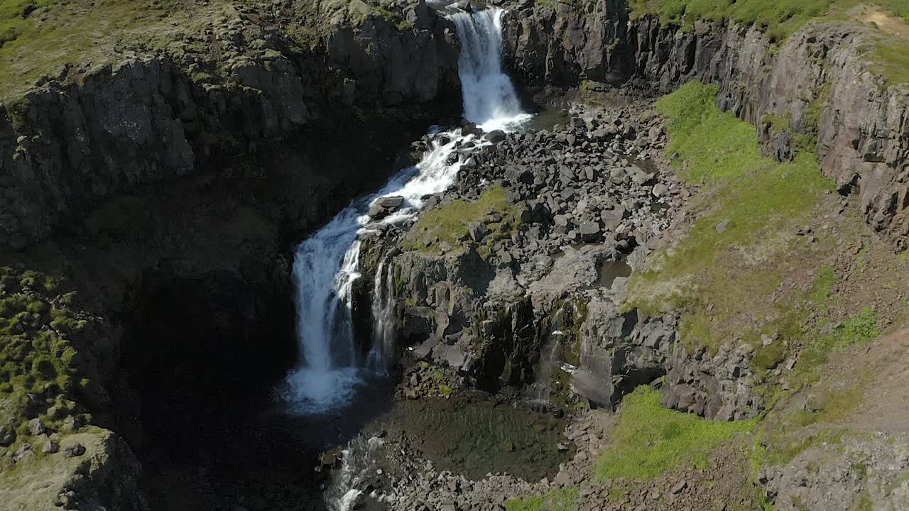Flight over Klifbrekkufossar waterfall with slow tilt upwards. Roaring and foaming icelandic waterfall with stony, mossy surroundings.