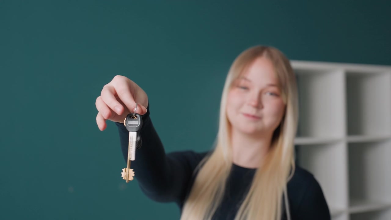 Donna caucasica che tiene nuove chiavi in un appartamento vuoto con parete color ottanio e scaffalature, capelli biondi, con maglione nero, sorridente mentre offre il portachiavi verso la telecamera, proprietaria di casa festante che celebra il trasloco