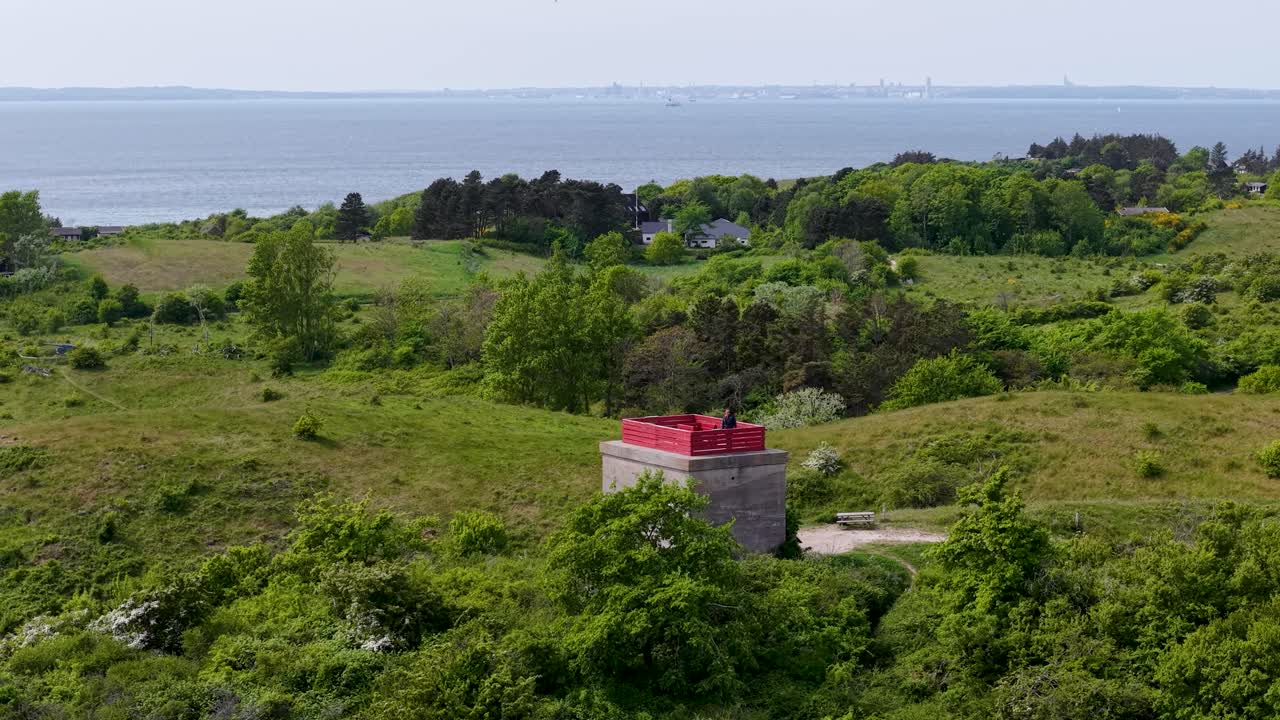 Aerial drone footage of Tyskertårnet, the German World War II bunker tower on the Danish coast, surrounded by green hills and overlooking the sea under cloudy daylight