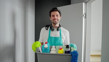 un limpiador moreno en una camiseta blanca, un delantal azul y auriculares inalámbricos negros sostiene un cuenco de plástico gris con herramientas de limpieza para la limpieza y detergentes en un apartamento moderno y brillante