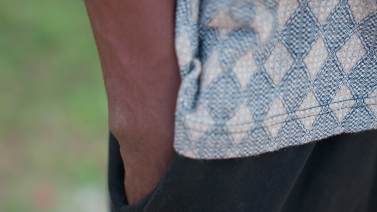 Nærbillede af sort shorts' lomme med hånd, kant af mønstret striktop, bar arm hviler i lommen, diskret bokeh-baggrund, afslappet holdning, casual sportstøjsstil, tekstur og synlige stingdetaljer.