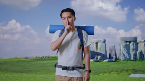 asian male hiker with mountaineering backpack looking at camera and making shh gesture while traveling in stonehenge