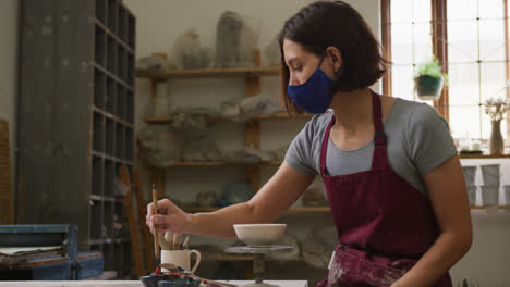 female potter wearing face mask and apron painting pot on potters wheel at pottery studio