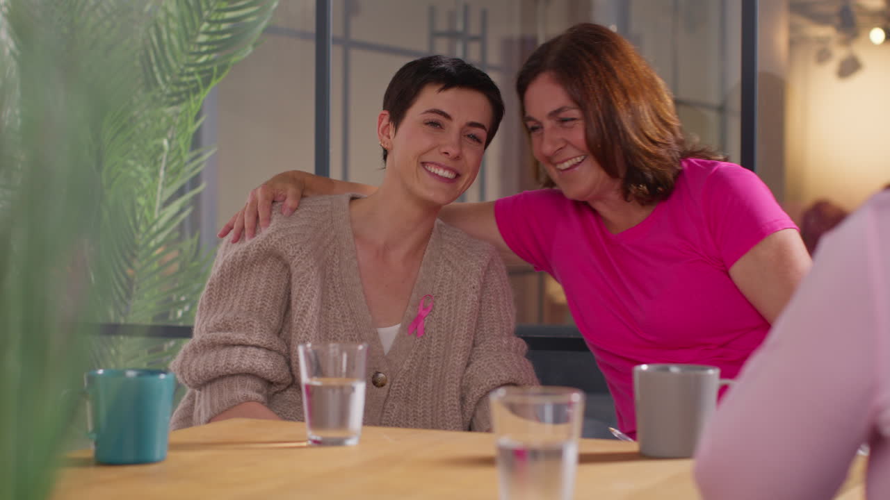 mujer con una cinta rosa de concienciación sobre el cáncer de mama siendo abrazada mientras habla en una reunión del grupo de apoyo de terapia para pacientes con tratamiento de cáncer