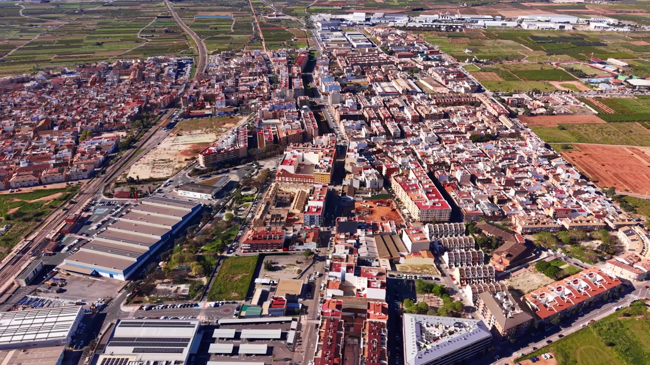 Vista aérea de Pucol, Valência, Espanha, mostrando densos blocos residenciais urbanos com telhados de terracota cercados por campos agrícolas e armazéns industriais