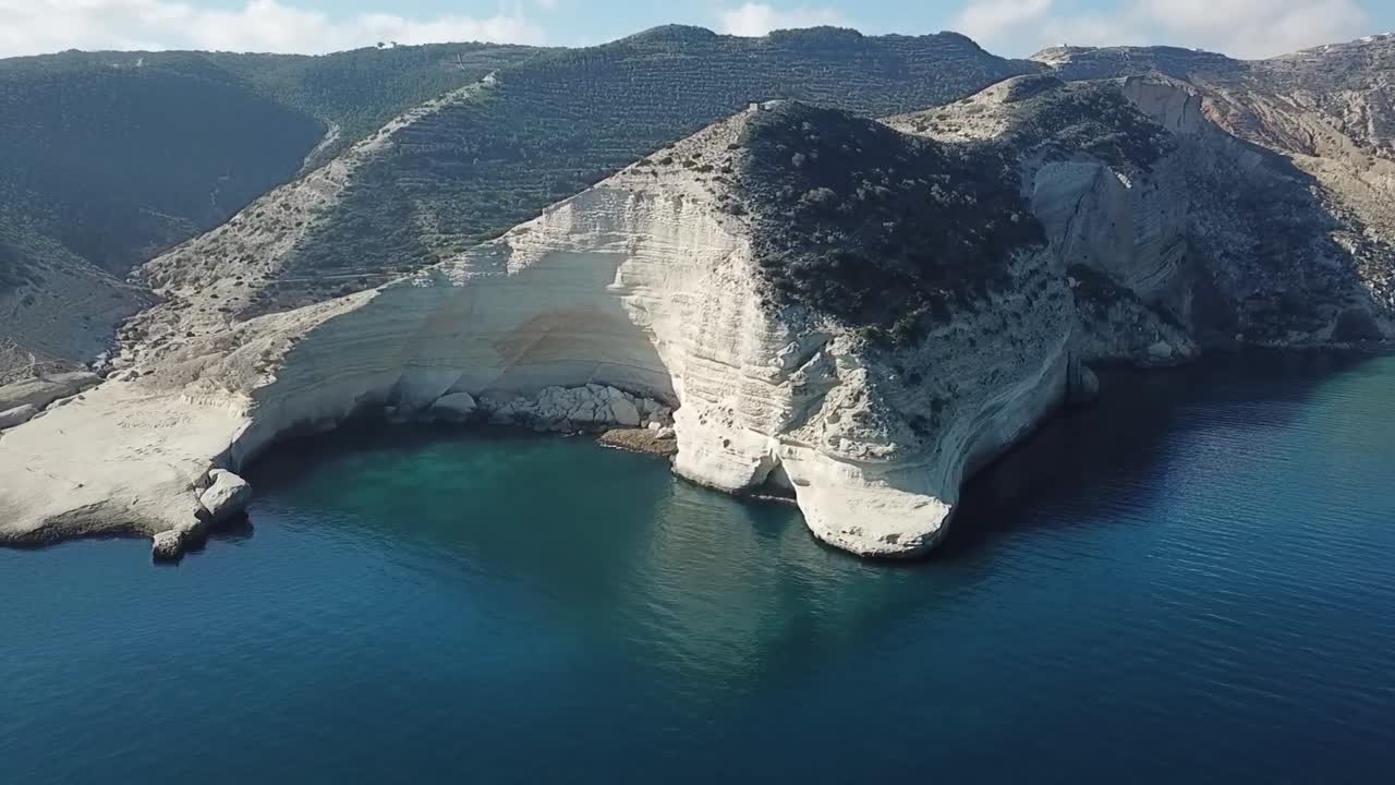 Wide drone shot capturing a small rocky island situated near a scenic coastline featuring a sandy beach and rugged mountains in the background