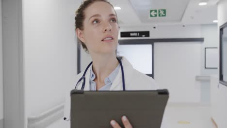 Happy-caucasian-female-doctor-walking-in-hospital-corridor-using-tablet,-in-slow-motion