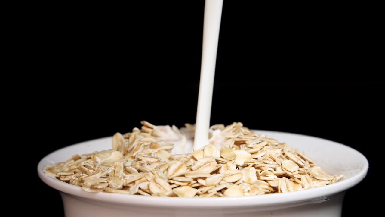 Milk is poured over oat cereal in a white bowl against a black background, creating a simple breakfast scene