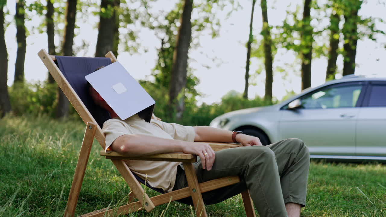 Man sits in the wood in a folding chair his face covered with laptop. Man smiles when the laptop descends. Car at backdrop.