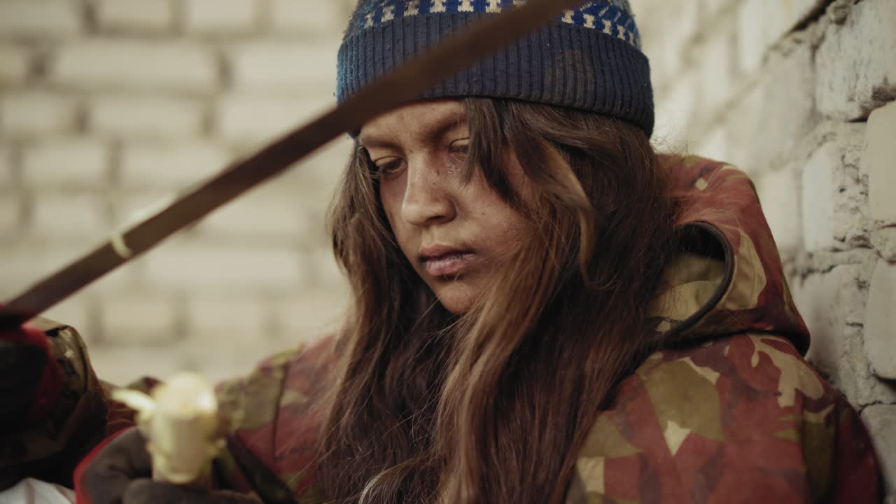 Close up of vagabond with dirty face wearing knit hat and gloves carving rough stick using knife while sitting against damaged brick wall, expression reflecting survival