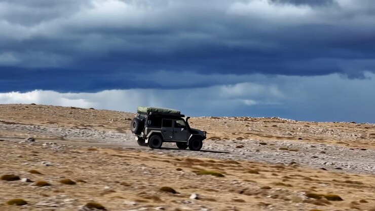 Jeep Crossing Arid Plateau Under Stormy Sky. Lone Expedition Vehicle With Roof Rack Kicking Dust Across Rugged