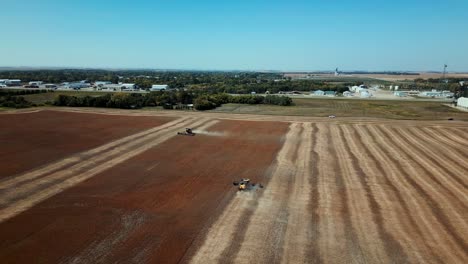 Un-Dron-De-Dos-Tractores-Combina-La-Agricultura-En-Un-Campo-Bajo-Un-Cielo-Azul-Claro-De-Verano-Con-Un-Pequeño-Pueblo-Detrás