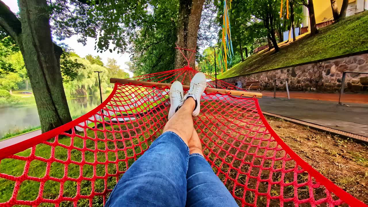 POV Relaxing in red hammock, swinging under trees next a pond with fountain in park