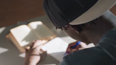 close rear view of student sitting at desk in library, writing notes in notebook with pen beside open book, focused on study as sunlight highlights pages creating calm atmosphere of learning