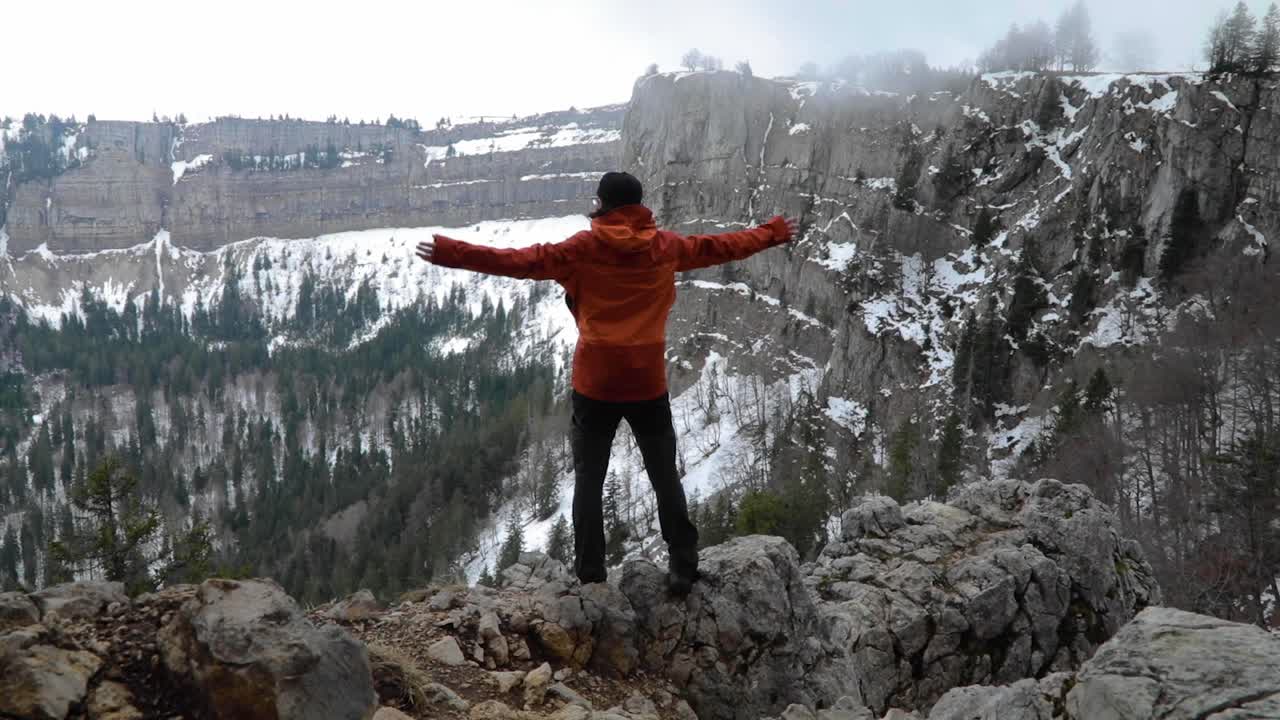 Hiker in winter clothes walk to cliff edge, raise arms. Overlook winter landscape. Success winning concept