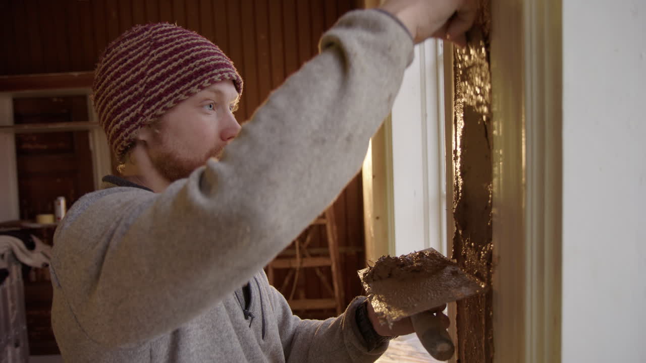 Nordic man backlit by floodlight applies clay plaster to house under renovation