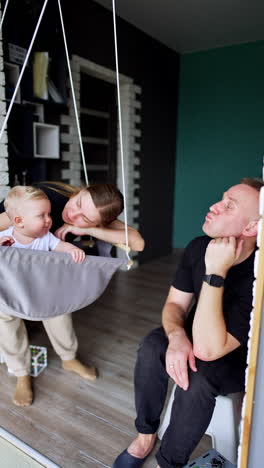 mother sways her lovely blond baby in a swing at home. father sits beside watching his child smiling. vertical video.